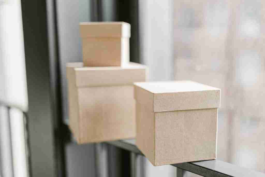 Tres cajas de cartón beige apiladas en distintos tamaños sobre un estante negro junto a una ventana