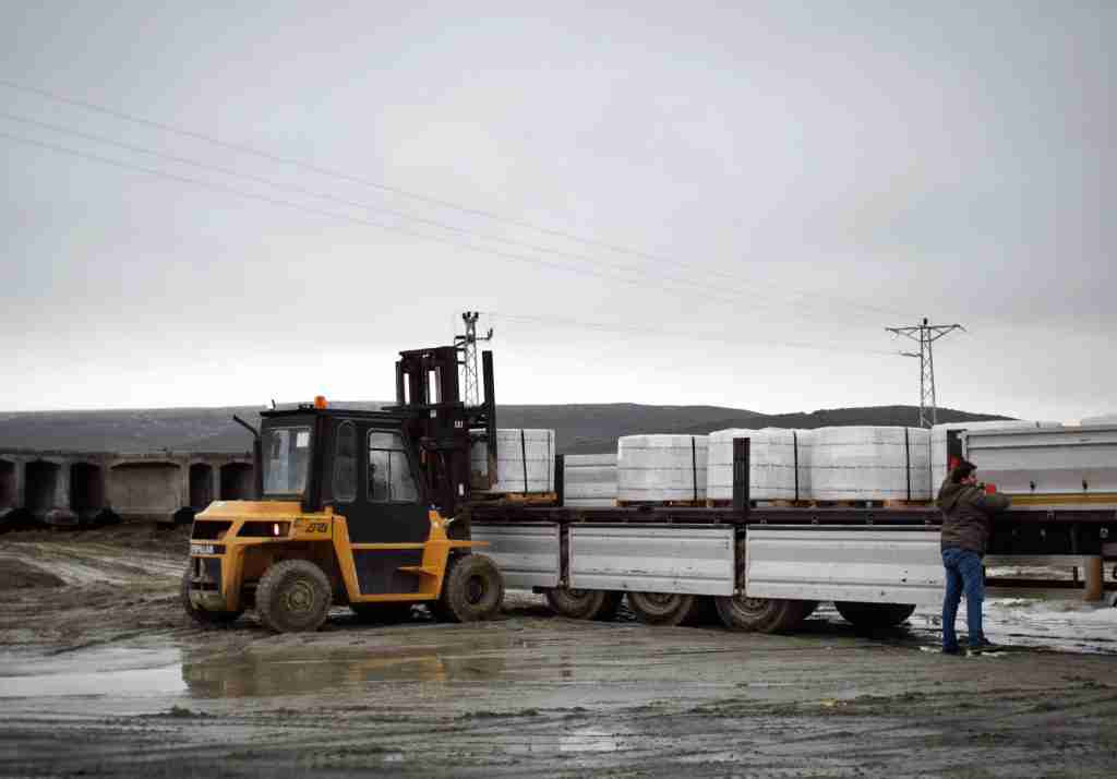 Carretilla elevadora Caterpillar amarilla cargando mercancía embalada en camión plataforma. Terreno embarrado y cielo nublado