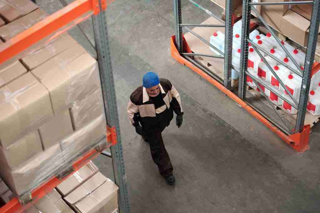 Operario con mono de trabajo y gorro azul caminando entre estanterías metálicas con cajas y bidones blancos en un almacén