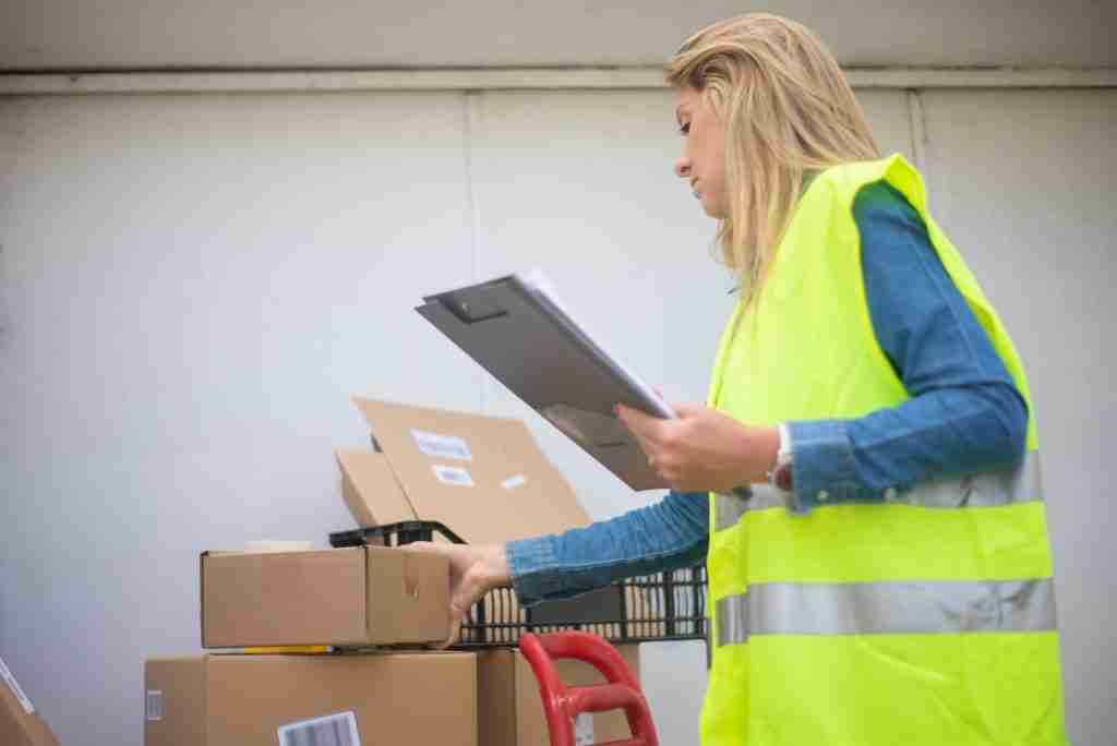 Mujer con chaleco reflectante amarillo revisa un portapapeles junto a varias cajas de cartón en un almacén